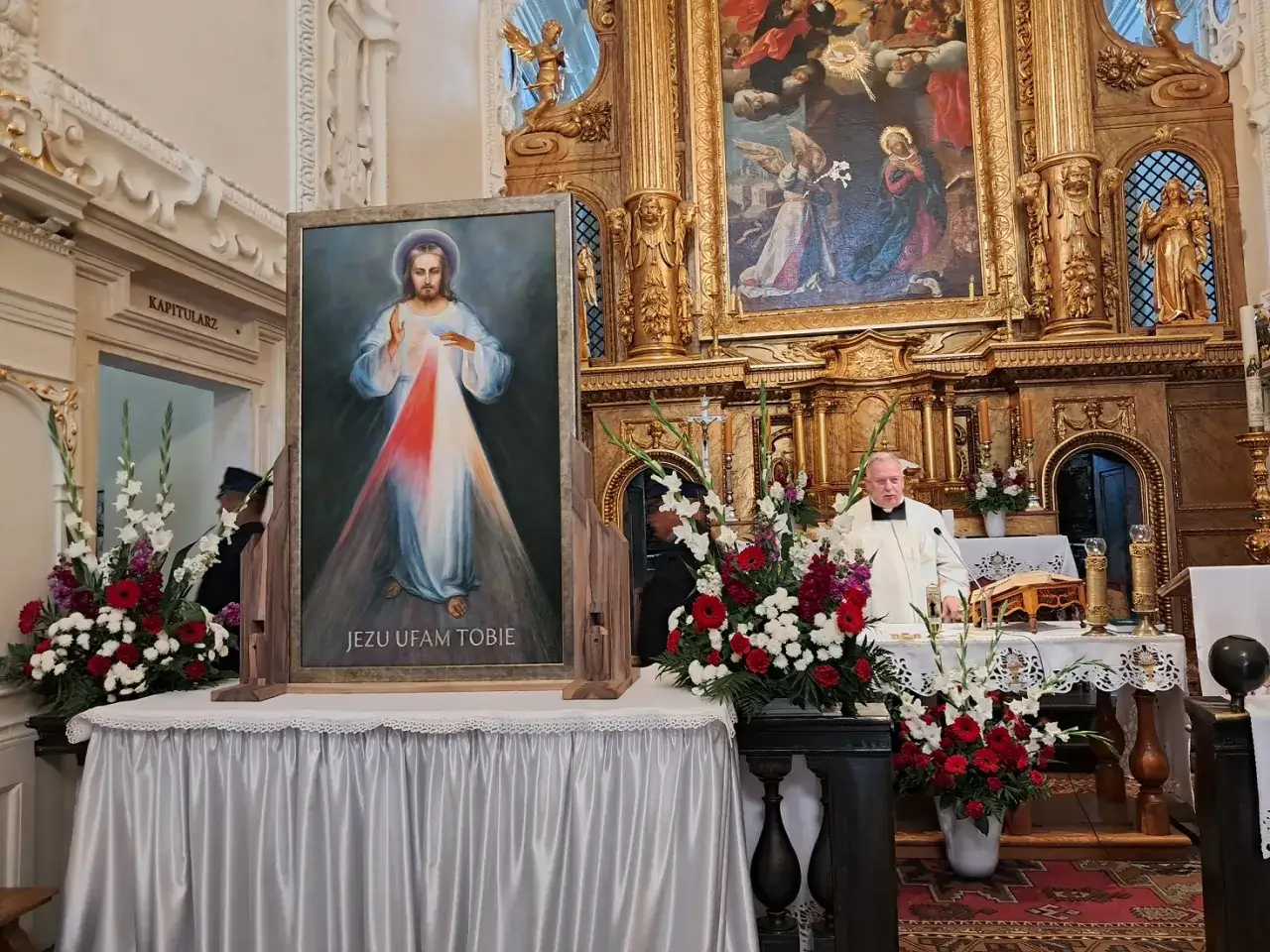 Obraz Jezusa Miłosiernego w kościele, gdzie kapłan odprawia mszę. Wokół kwiaty, symbolizujące piękno życia, ale też samotność w obliczu wiary.