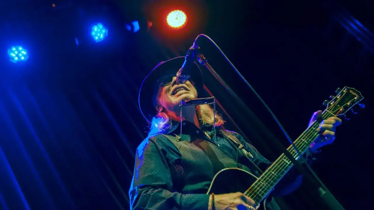 Elliott Murphy en Zaragoza, tocando la guitarra y la armónica bajo luces azules y rojas.