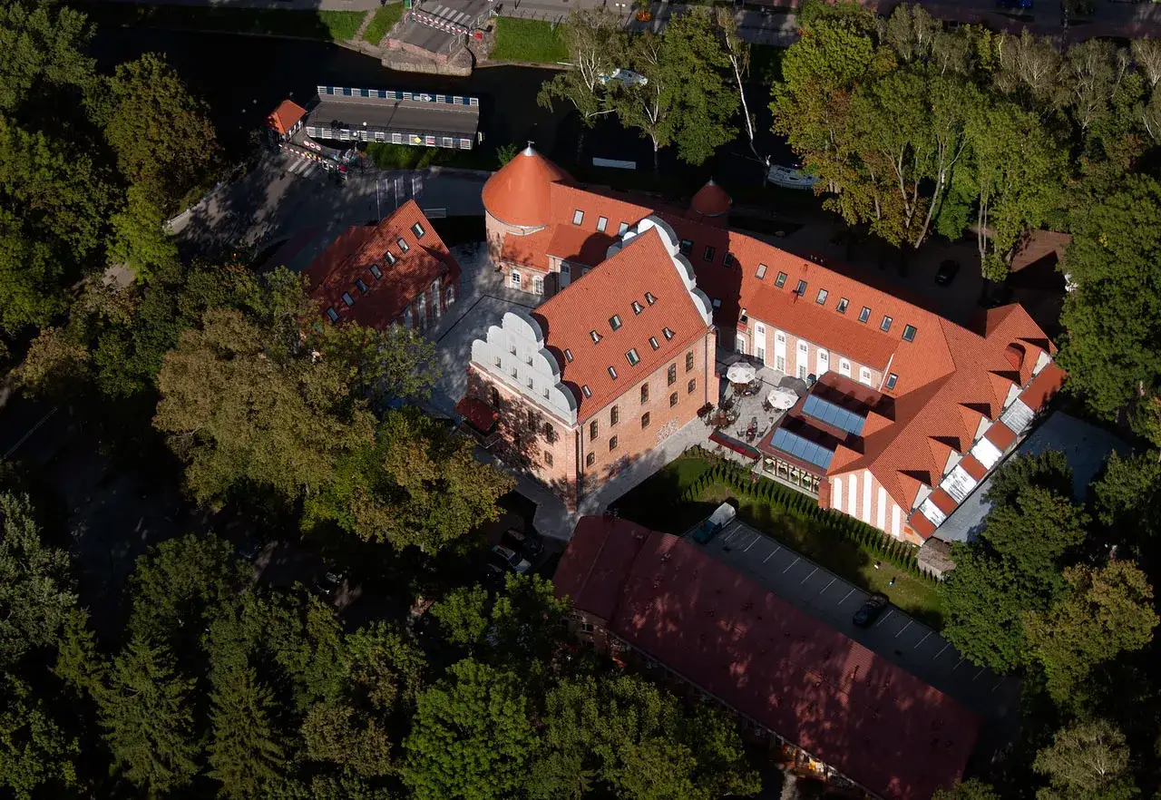 Widok z lotu ptaka na zamek w Giżycku otoczony zielenią. Czerwone dachy i ceglane mury tworzą malowniczy krajobraz.