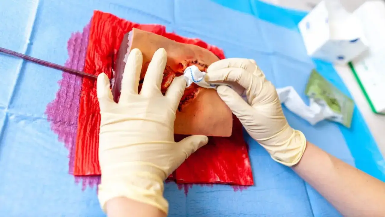 Ręce w rękawiczkach ćwiczą użycie środk&oacute;w hemostatycznych na symulatorze rany. Czerwona gaza nasiąknięta płynem imitującym krew.
