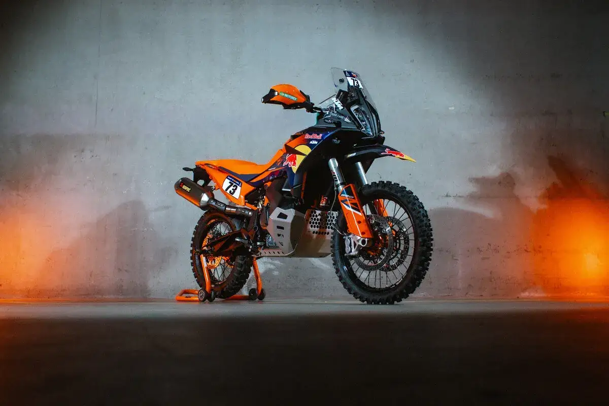 La KTM 490 Adventure, lista para la acci&oacute;n, con su distintivo naranja y azul, destaca sobre un fondo texturizado con luces c&aacute;lidas.