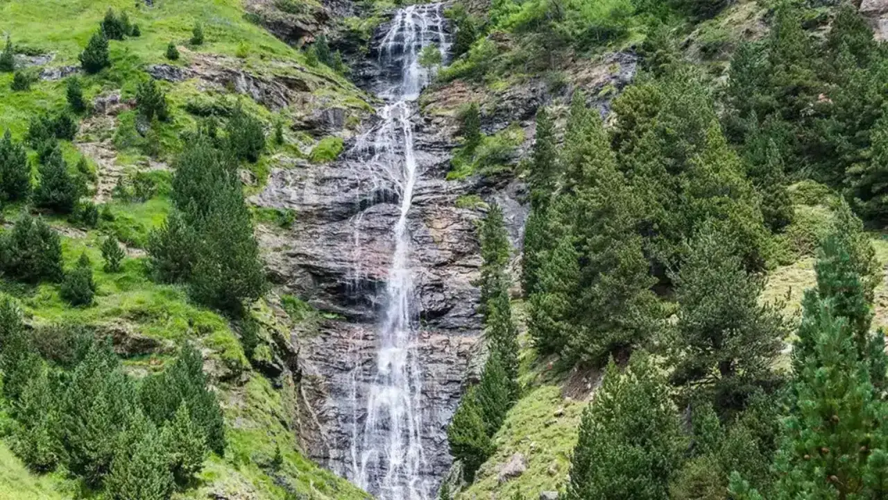 Cascada en los Pirineos, rodeada de pinos y vegetaci&oacute;n verde. El agua desciende por rocas escarpadas, alimentando quiz&aacute;s lagos ocultos en el valle.