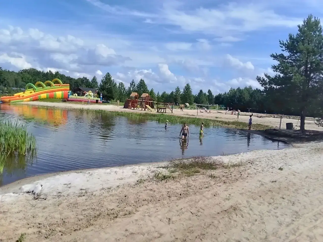 Sielska pogoda nad wodą. Dzieci kąpią się, a w tle zjeżdżalnia dmuchana i plac zabaw.