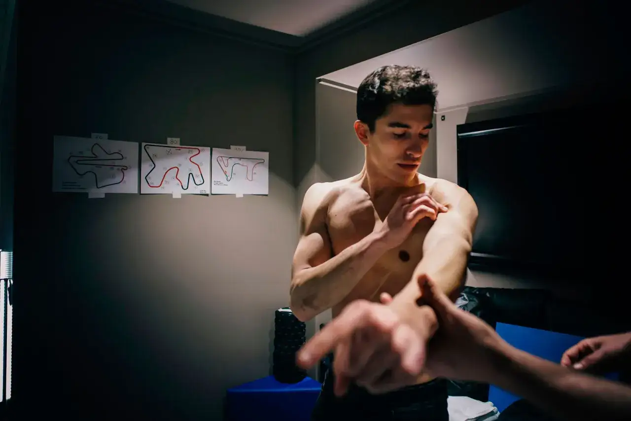 Marc M&aacute;rquez se examina el brazo, quiz&aacute;s recuper&aacute;ndose de una lesi&oacute;n, con diagramas de circuitos de carreras en la pared.
