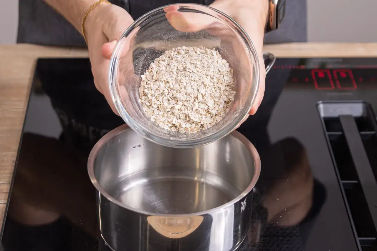 Haferschleim rezept: Haferflocken werden aus einer Schüssel in einen Topf mit Wasser gegossen.