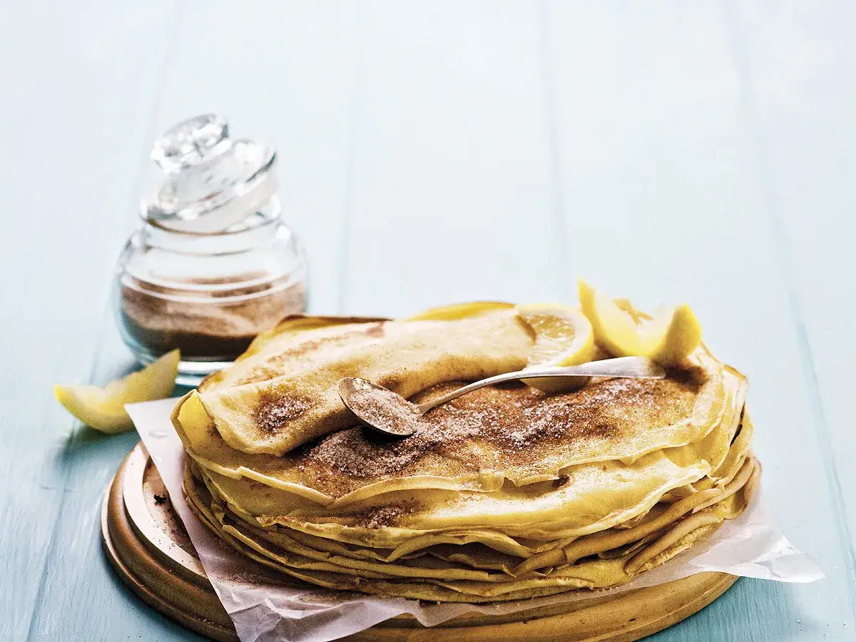 Pile de crêpes au lait entier, saupoudrées de sucre et de cannelle, garnies de quartiers de citron.