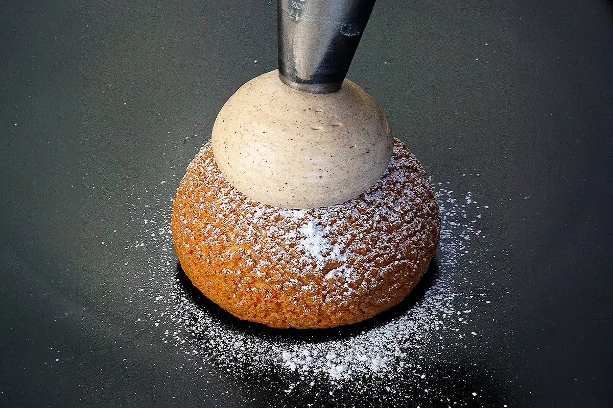 Une poche à douille dépose une généreuse boule de ganache montée chocolat sur un chou saupoudré de sucre glace.