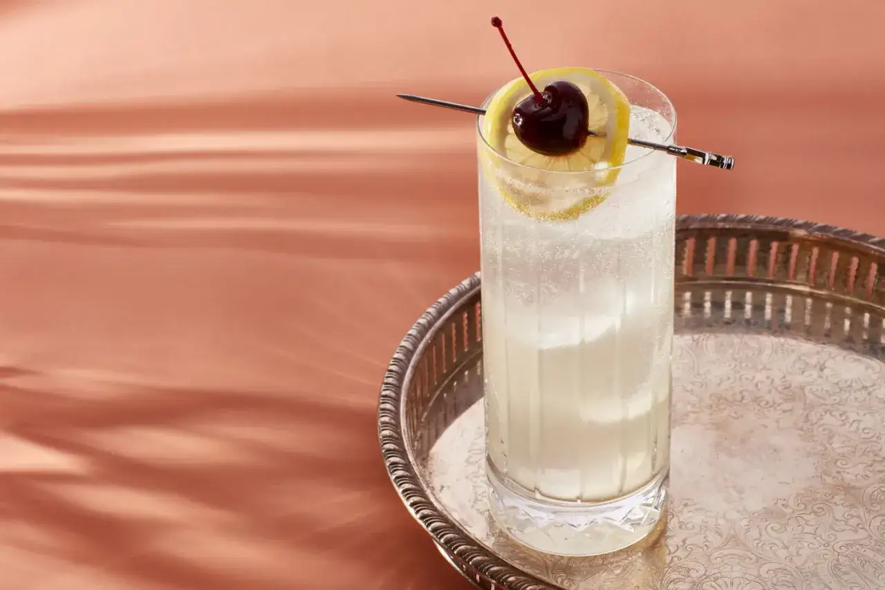 En h&ouml;g, svalkande Tom Collins med citronskiva och k&ouml;rsb&auml;r, serverad p&aring; ett silverfat.