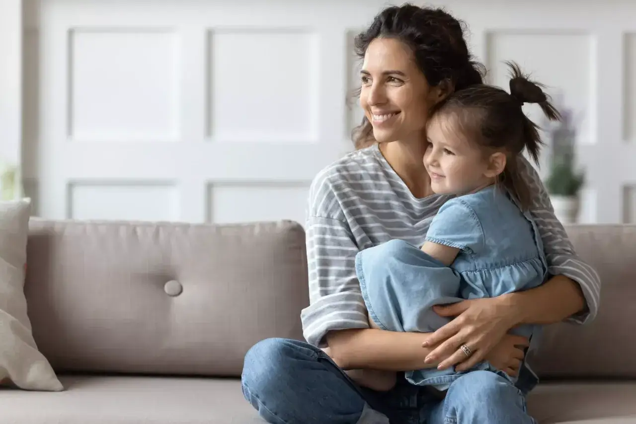 Uśmiechnięta mama tuli córkę, wspierając jej rozwój emocjonalny dziecka. Siedzą na kanapie, patrząc w dal.