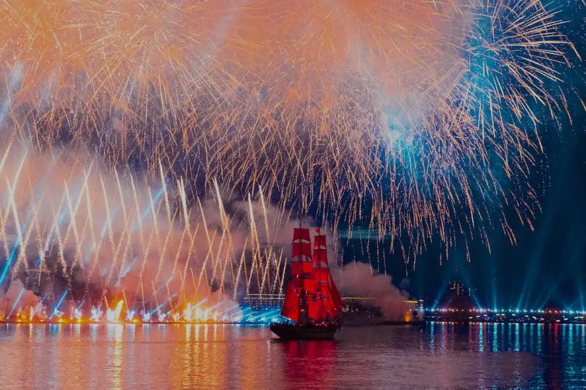 Spektakularny pokaz pirotechniczny rozświetla nocne niebo nad wodą, gdzie płynie żaglowiec z czerwonymi żaglami.