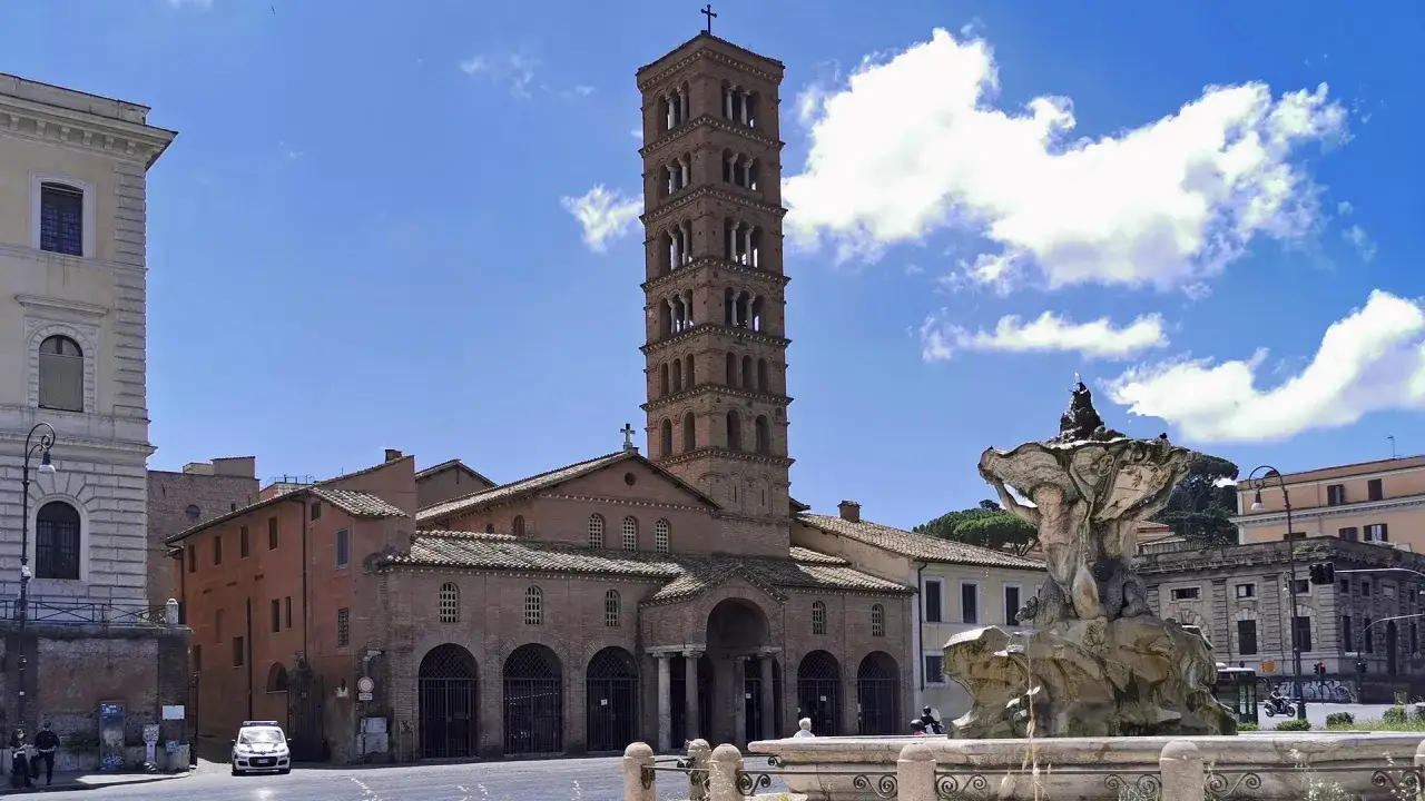 Fontana con la **Boca de la Verita** e la Basilica di Santa Maria in Cosmedin a Roma, sotto un cielo azzurro con nuvole bianche.