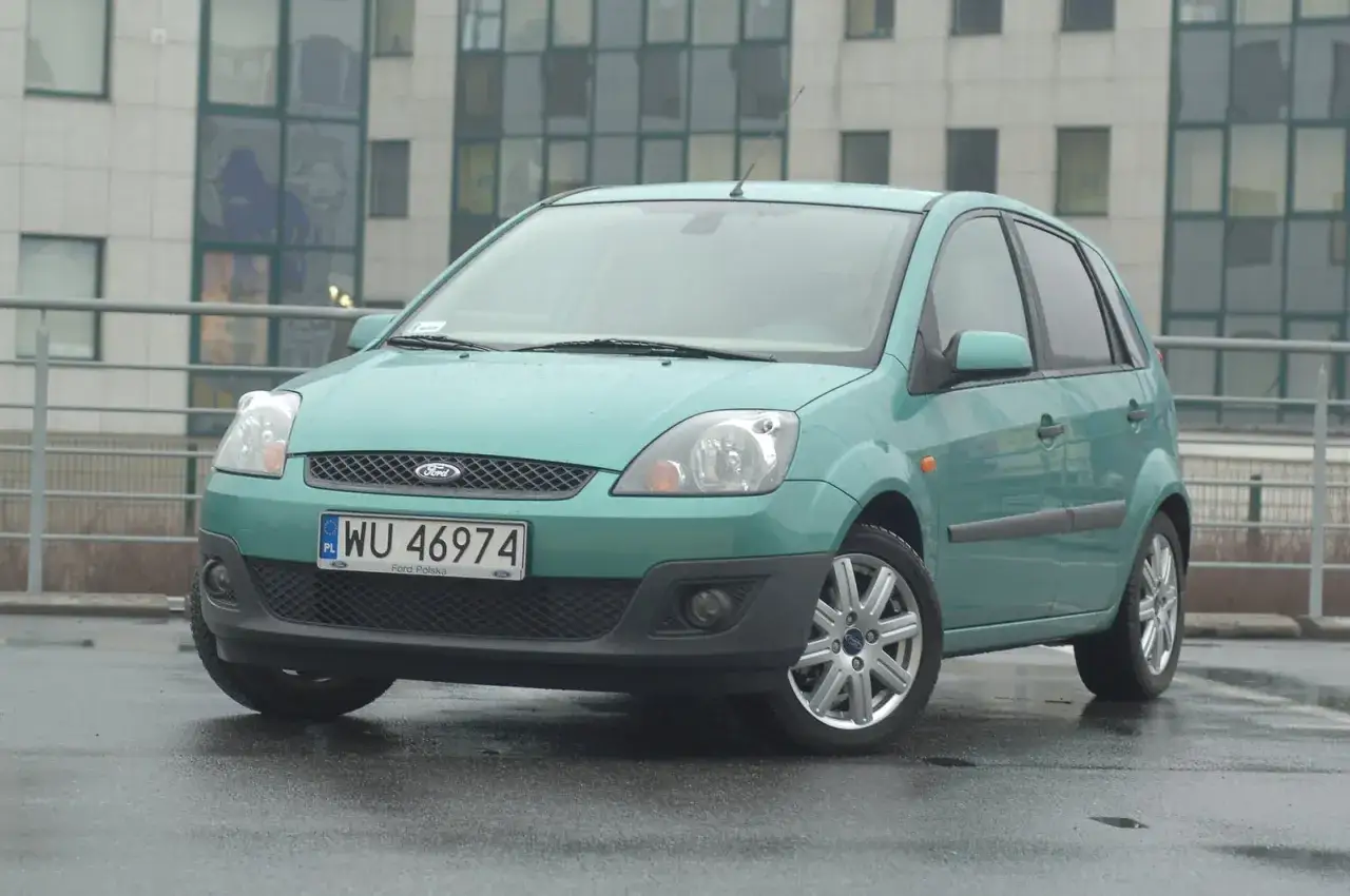 Zielony Ford Fiesta MK5 z polskimi tablicami WU 46974 stoi na mokrym parkingu przed nowoczesnym budynkiem.