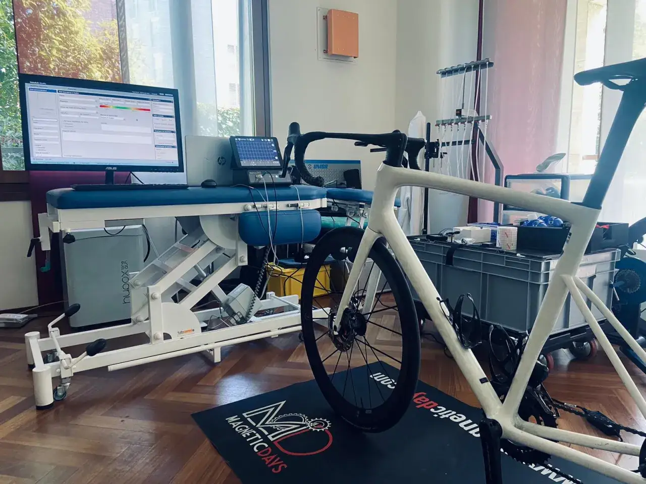 Bicicletta da corsa bianca su rullo, con monitor che mostra dati di allenamento e grafici, forse per misurare l'acido lattico.