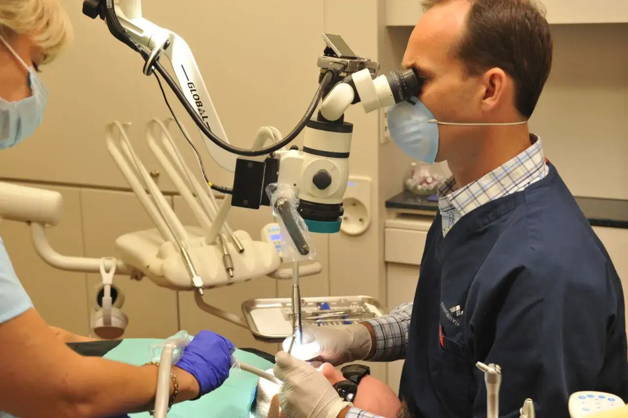 Dentysta pod mikroskopem wykonuje powtórne leczenie kanałowe. Asystentka przygotowuje narzędzia.