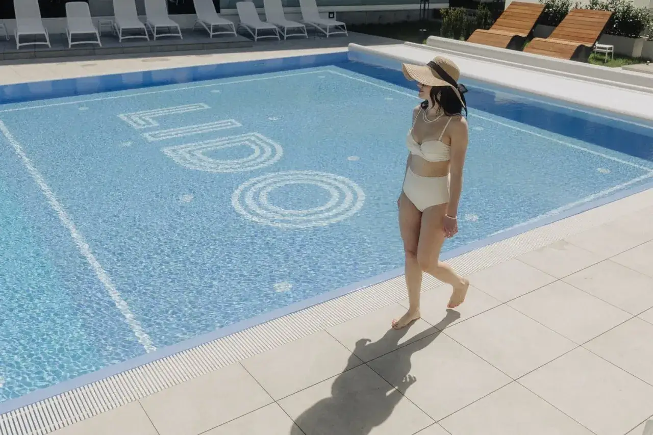 Kobieta w stroju kąpielowym i kapeluszu spaceruje przy basenie z napisem LIDO. Idealne miejsce na wakacje w Jurata hotel Lido.