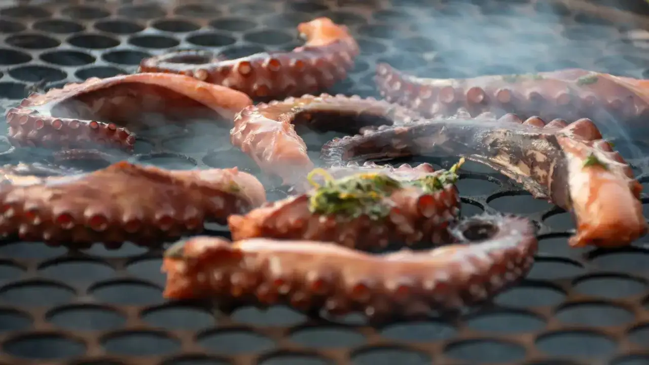Tent&aacute;culos de pulpo a la brasa humeantes sobre una parrilla, con hierbas verdes espolvoreadas.