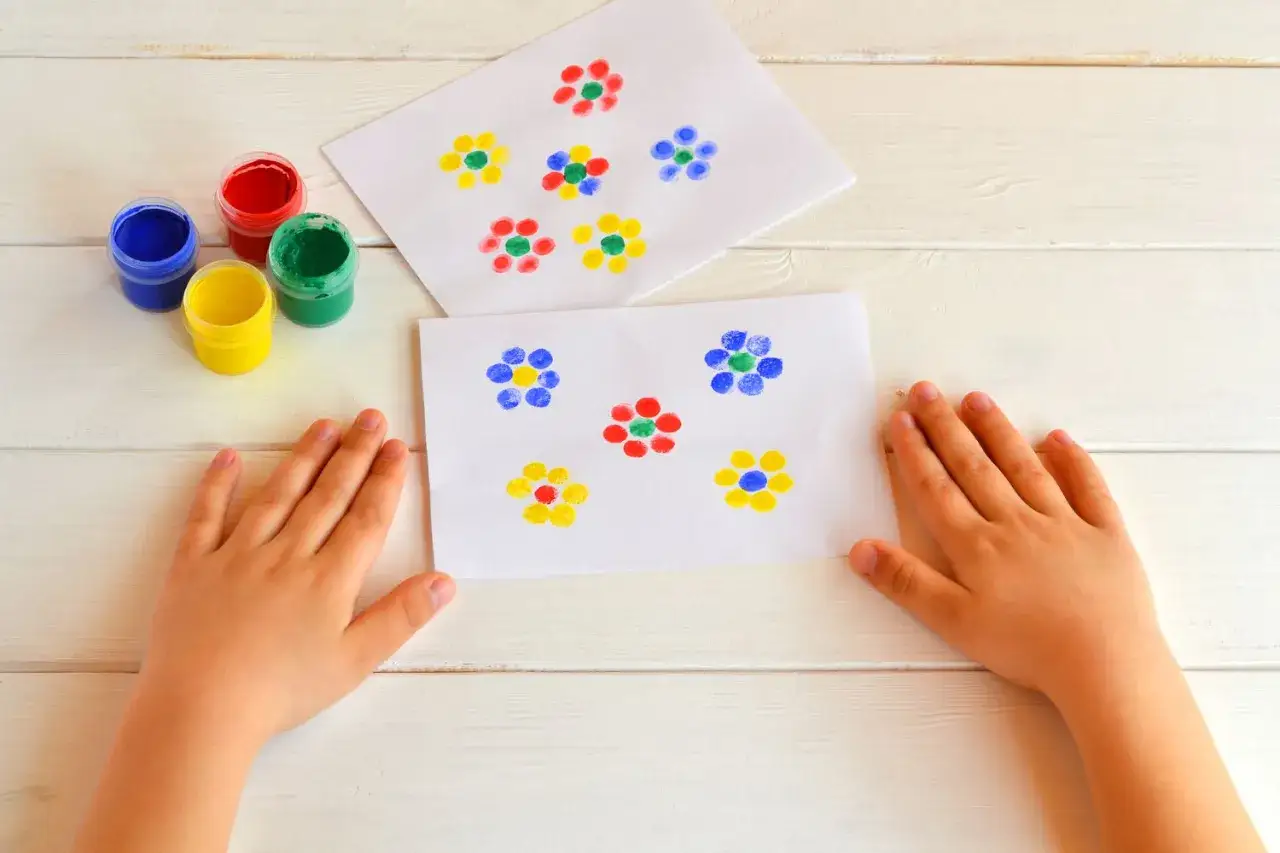 Manos de ni&ntilde;o creando flores con pintura usando la t&eacute;cnica de huella dedo. Hay botes de pintura azul, roja, verde y amarilla.