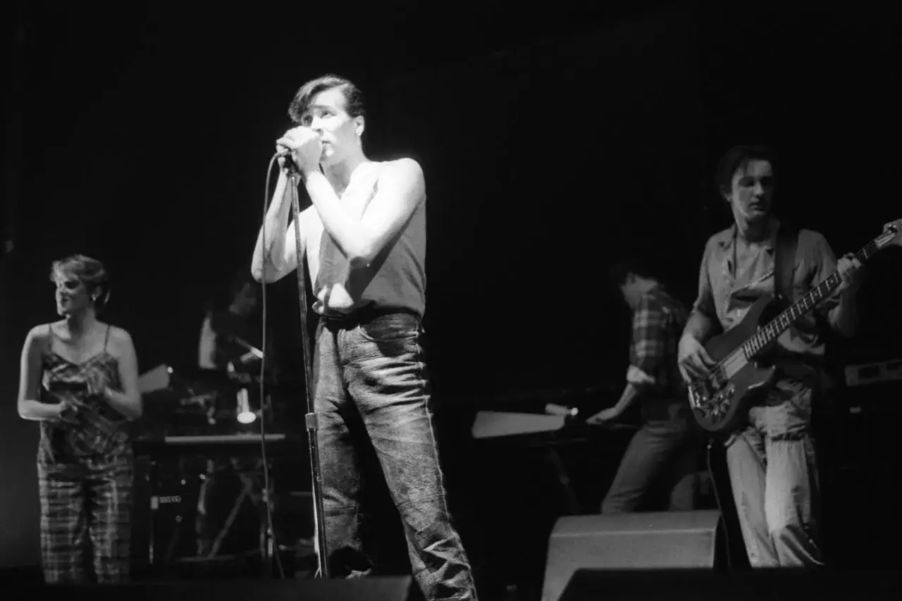 Un groupe rock anglais des années 80 en pleine performance. Le chanteur, au premier plan, tient un micro. À ses côtés, un bassiste et d'autres musiciens sur scène.