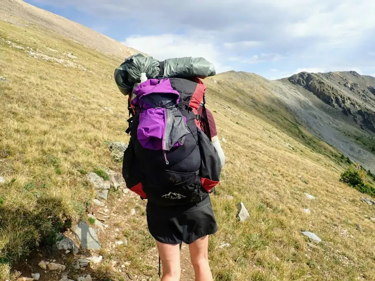 Persona con mochila mountain, lista para la aventura en la ladera de una montaña con hierba seca y cielo nublado.