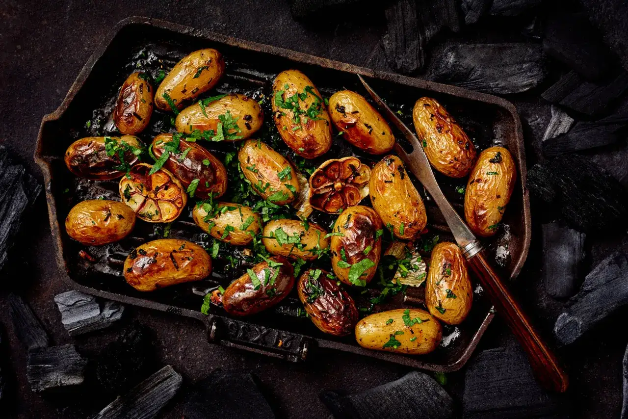 Små, gyllene potatisar med örter i en grillpanna, perfekta som potatis till grillat. Kolbitar ramar in scenen.