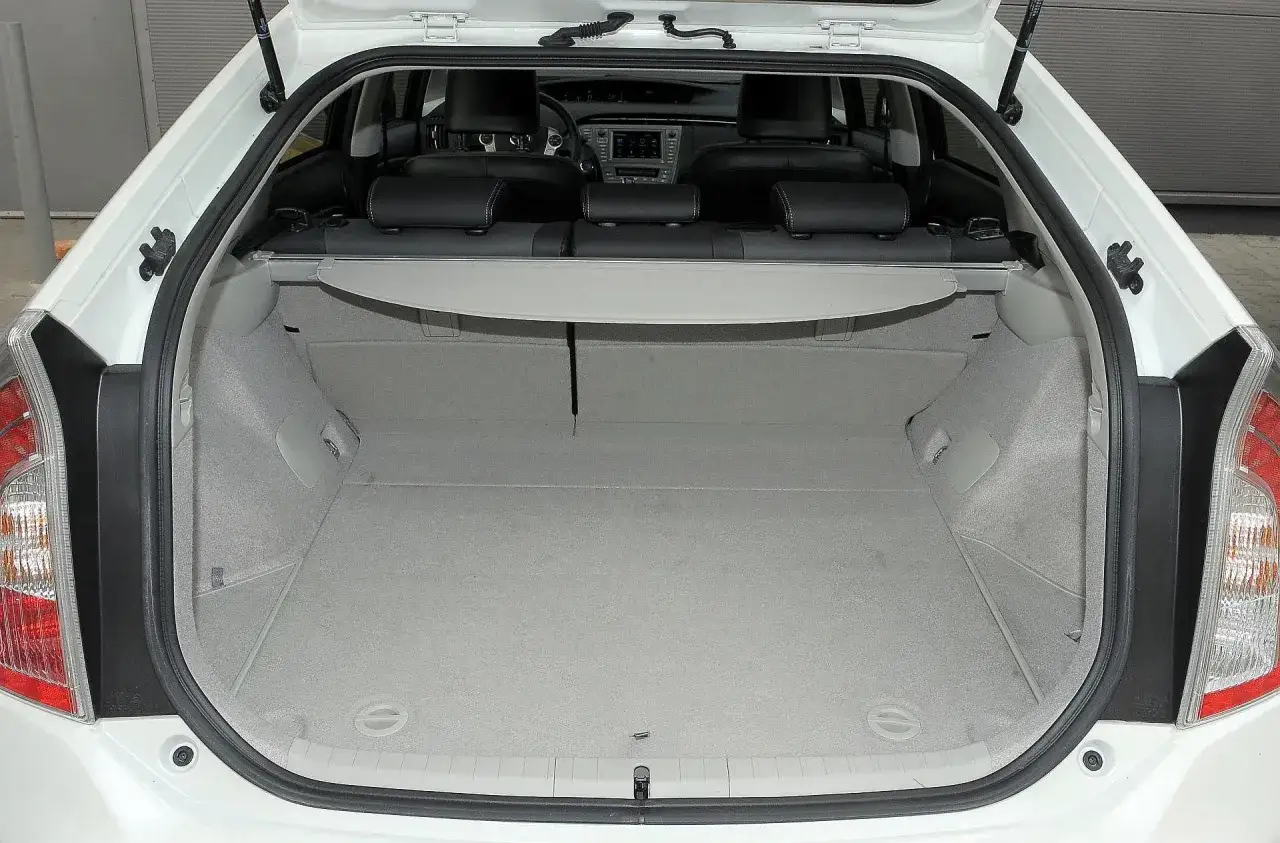 Otwarty bagażnik białego **Toyoty Prius 2** z jasną wykładziną i zasłonką. Widać tylne siedzenia i deskę rozdzielczą.