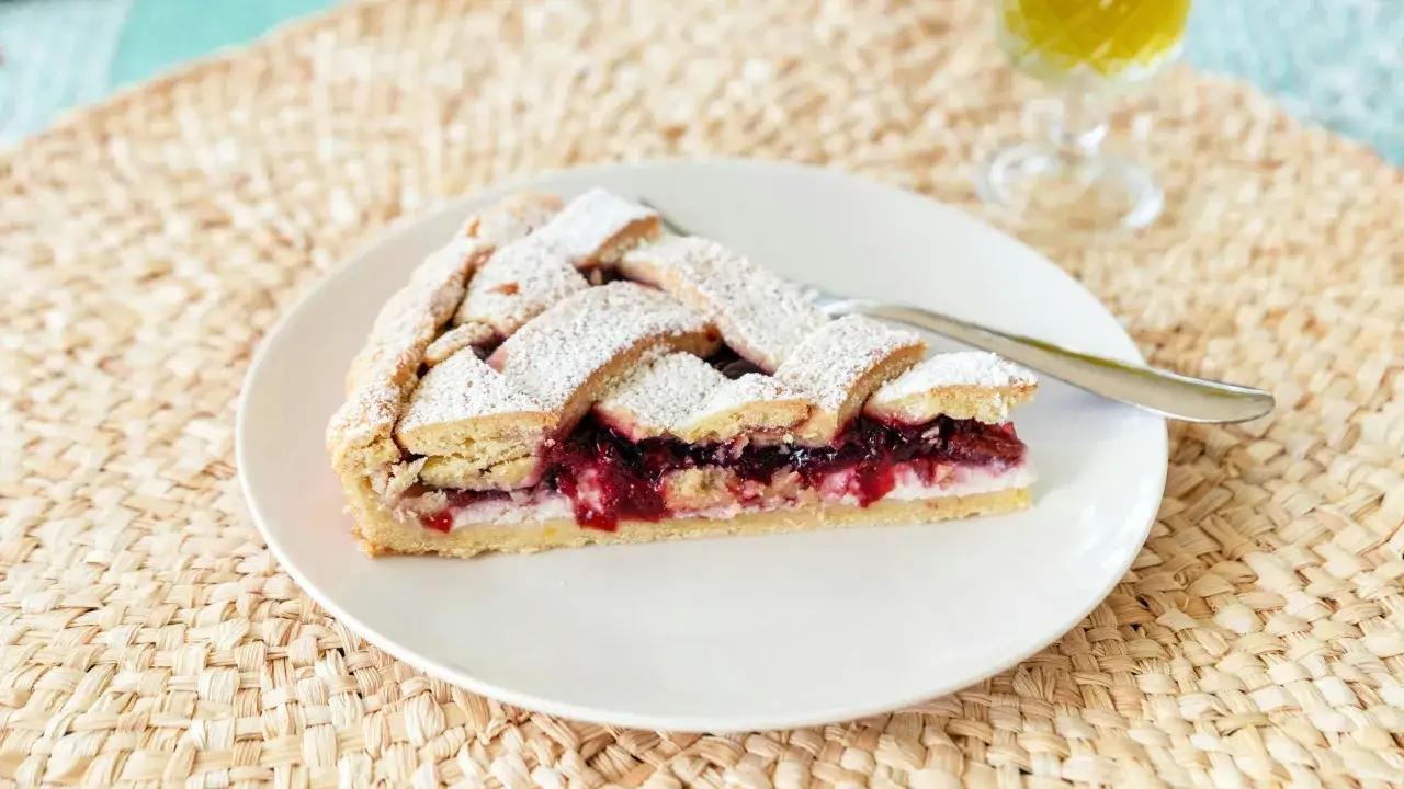 Una fetta di crostata di ricotta con confettura di ciliegie, cosparsa di zucchero a velo, su un piatto bianco.