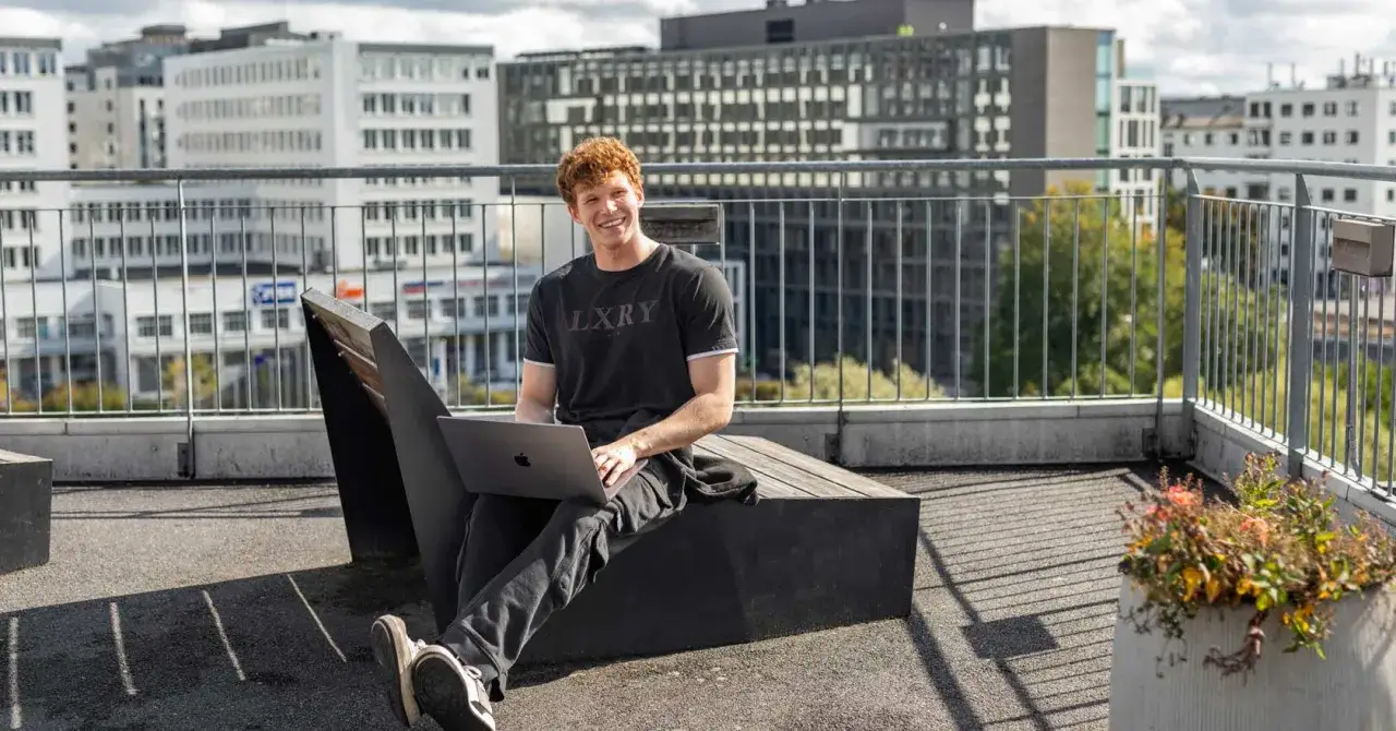 Uśmiechnięty student z rudymi włosami pracuje na laptopie na dachu budynku, z widokiem na nowoczesną architekturę, przypominającą kampus uniwersytetu w Oslo.