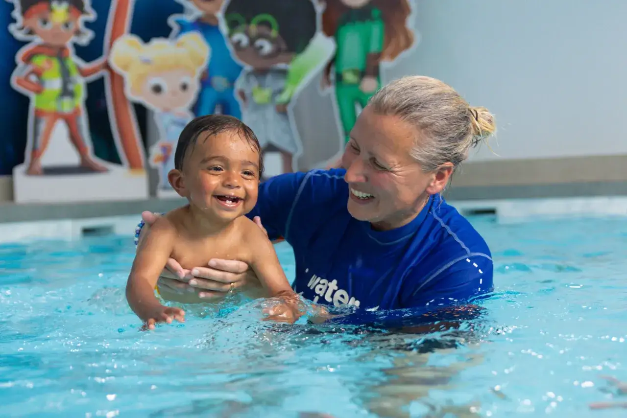 A smiling swim instructor holds a happy baby in a pool. This scene makes you wonder how much do swim instructors make for such rewarding work.