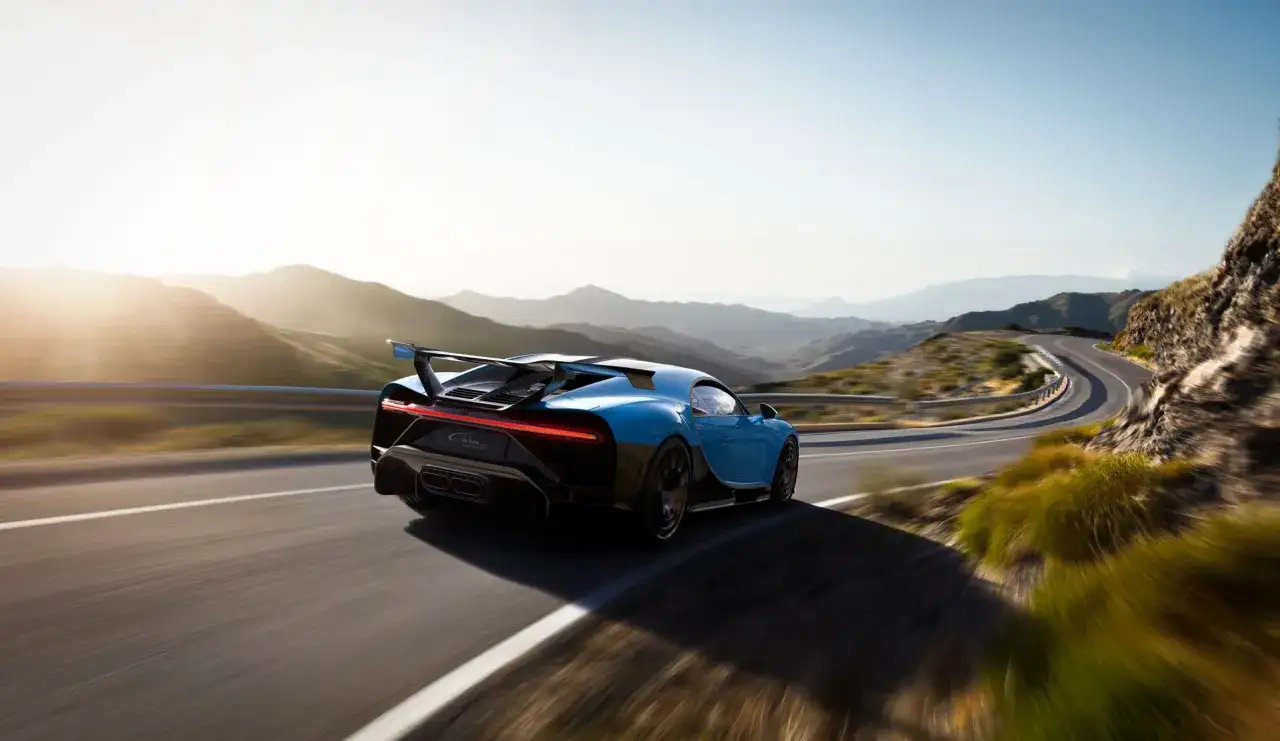Blauer Chiron Pur Sport rast auf kurviger Bergstraße, die Sonne scheint.
