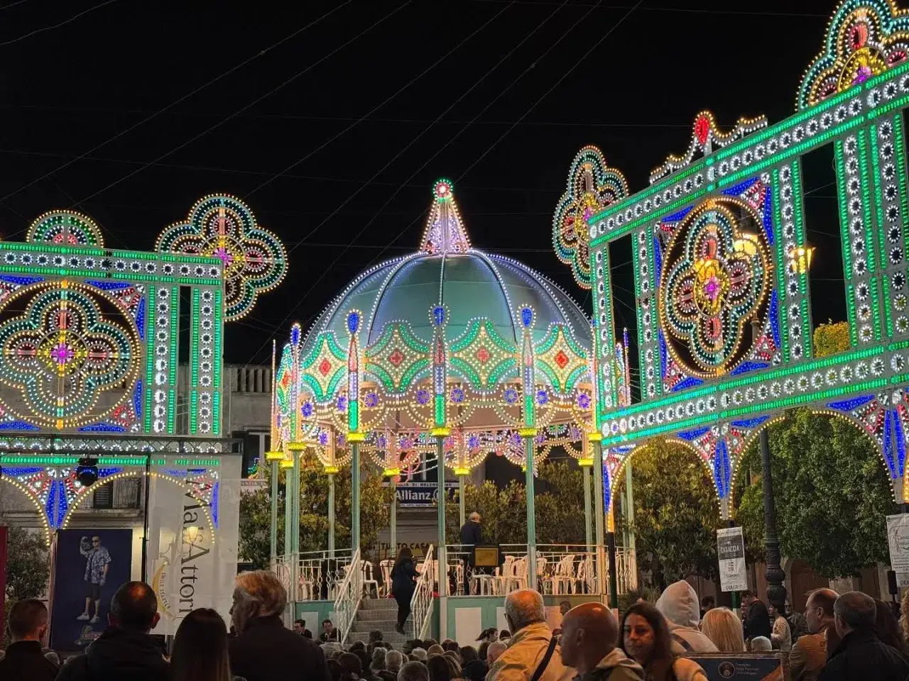 Luci scintillanti illuminano una piazza pugliese, creando un'atmosfera magica per gli eventi stasera in Puglia. Un palco centrale e decorazioni elaborate invitano alla festa.