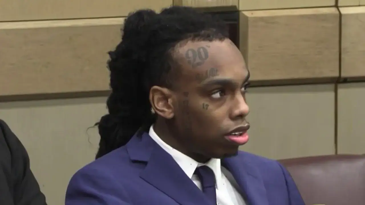 YNW Melly w garniturze, z tatuażami na twarzy, siedzi w sądzie.