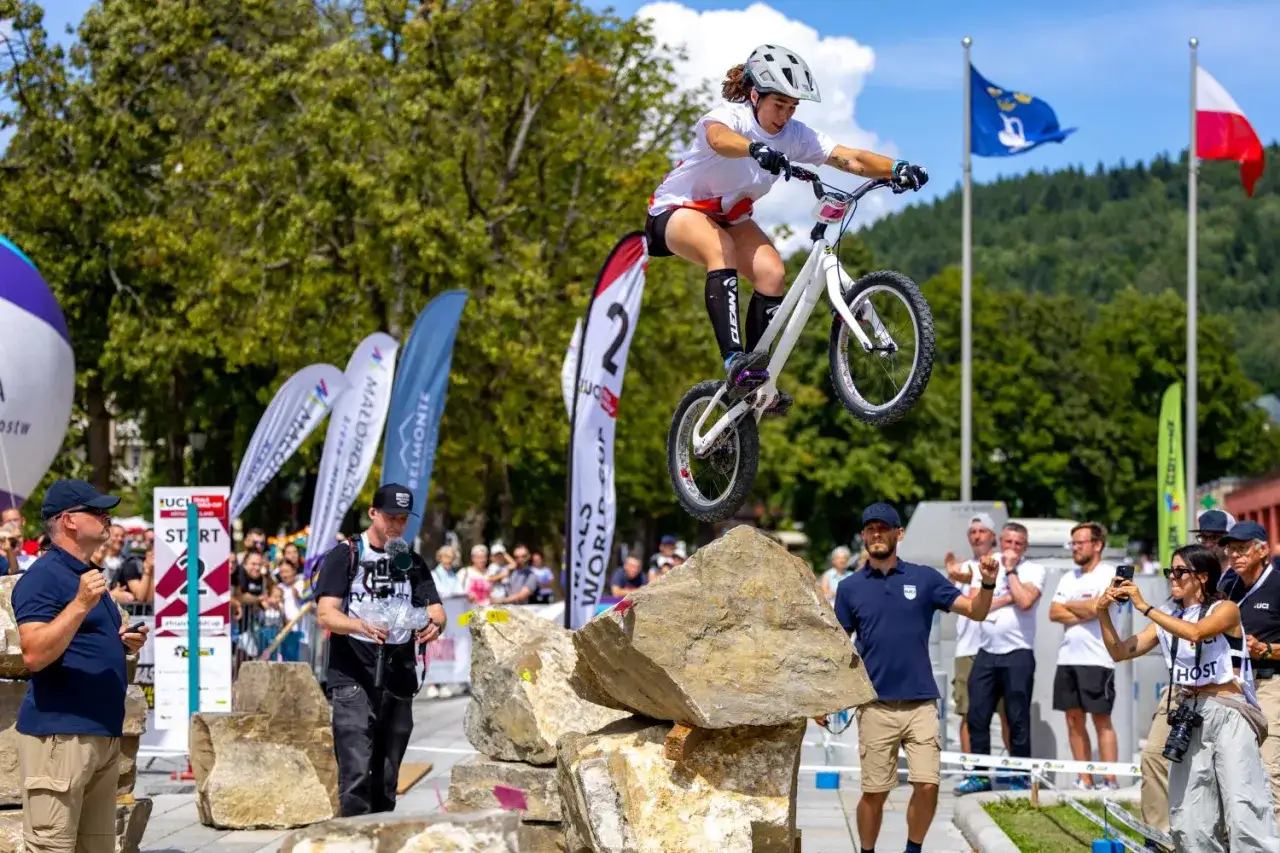 Kobieta w kasku wykonuje skok na rowerze podczas zawodów trialowych. To widowiskowy sport, gdzie liczy się precyzja i równowaga.