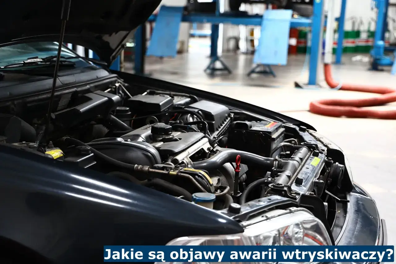 Silnik samochodu z otwartą maską. Widać elementy układu paliwowego, akumulator i przewody. Tekst na dole: 