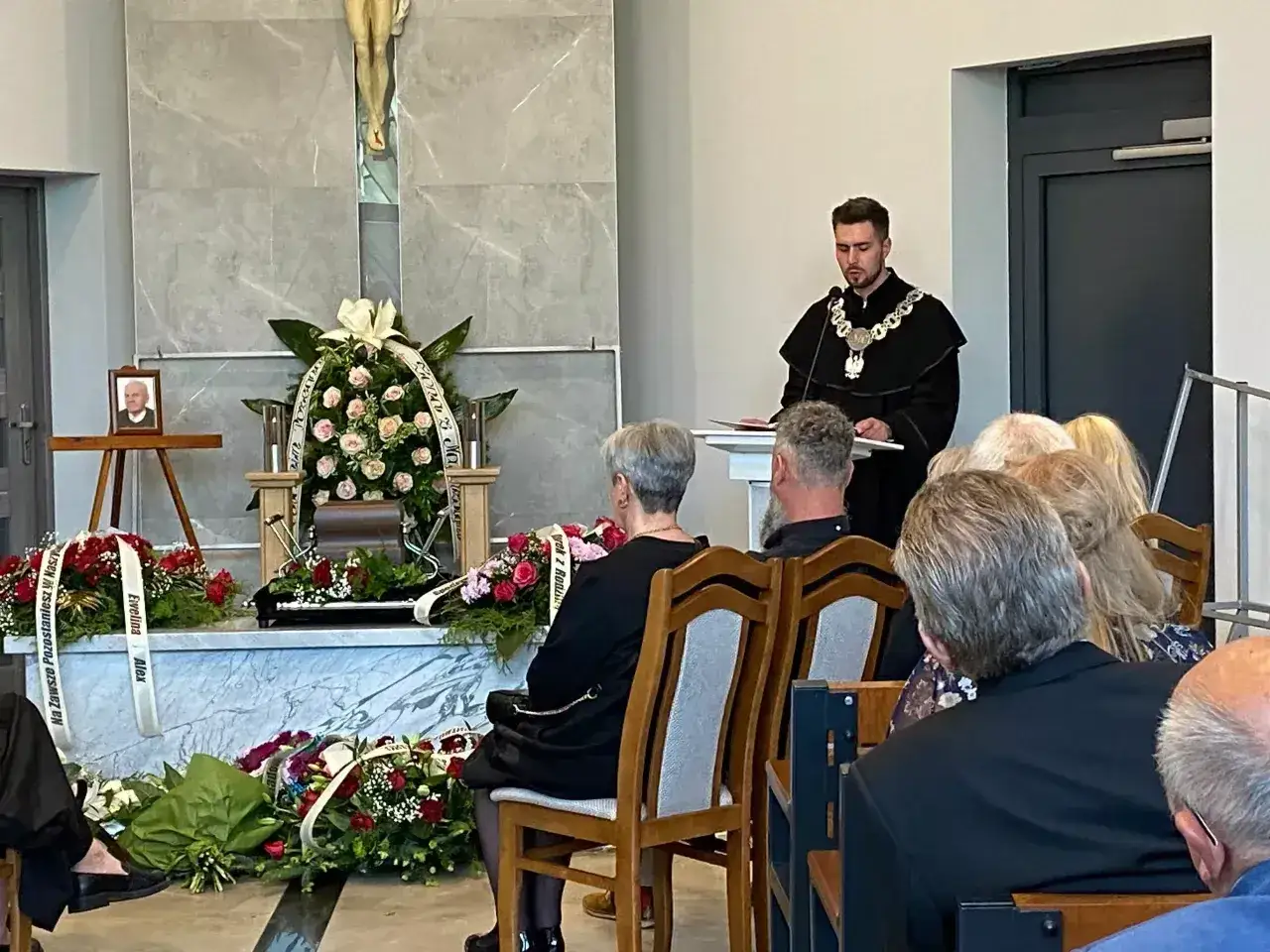 Uroczystość świecki pogrzeb. W sali zgromadzeni żałobnicy, przed nimi trumna ozdobiona kwiatami. Mężczyzna w todze wygłasza mowę.