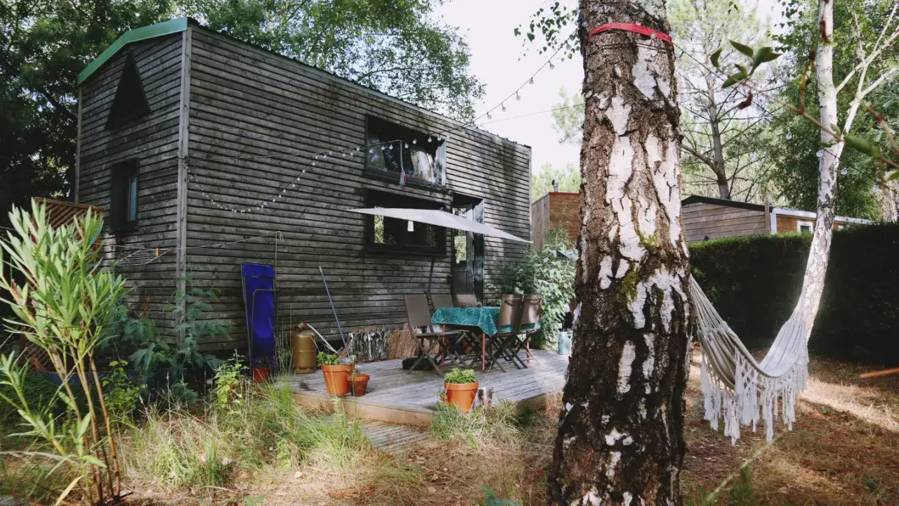 Une tiny house en bois dans les Landes, avec une terrasse, une table et des chaises, un parasol, et un hamac suspendu entre deux arbres.