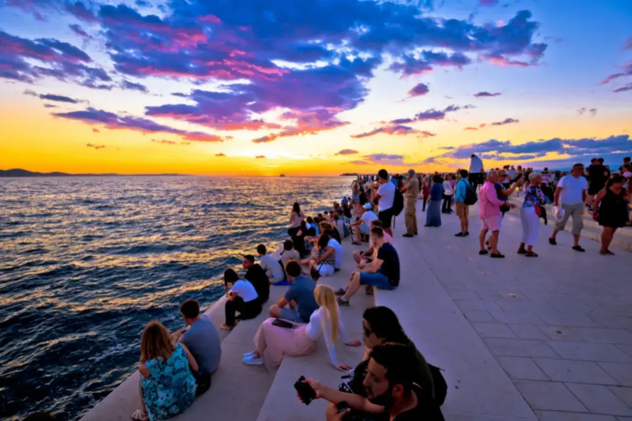 Gente sentada en el paseo mar&iacute;timo de Zadar, disfrutando de una espectacular puesta de sol sobre el mar Adri&aacute;tico. Un momento perfecto para ver en Zadar.