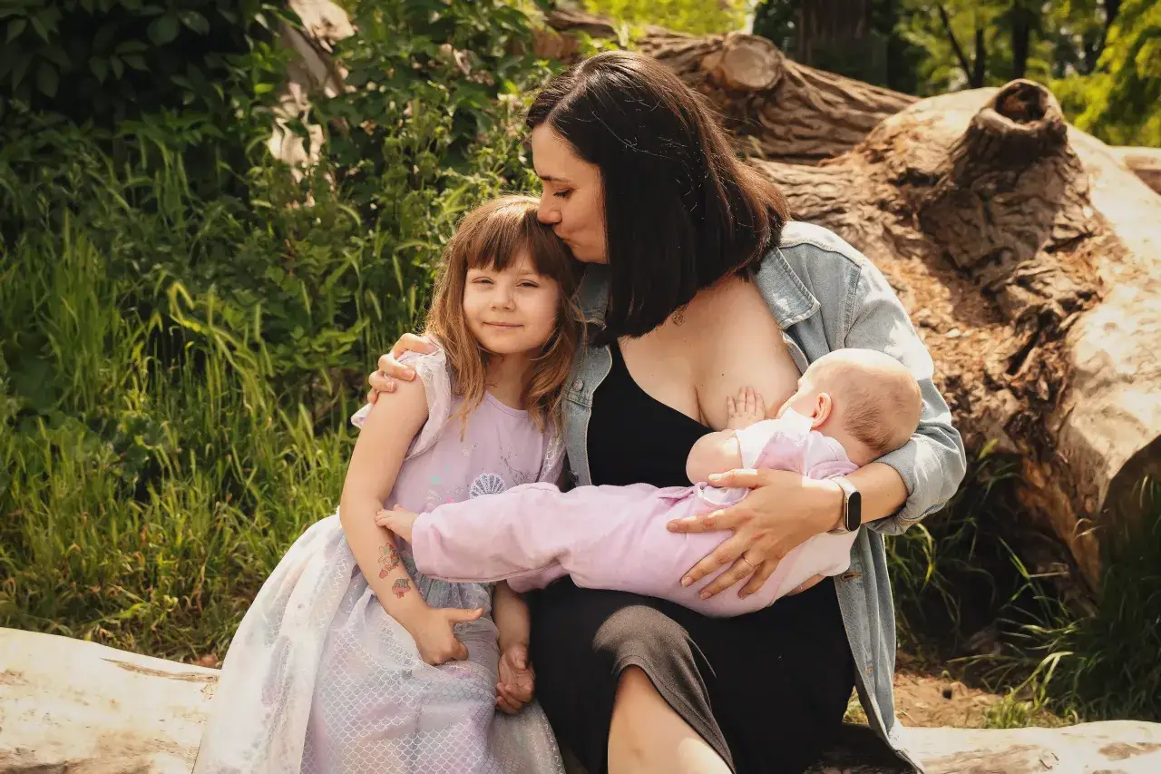 Mama karmi piersią maluszka, podczas gdy starsza córka w sukience z syrenką przytula się do niej. Obrazek idealny na światowy tydzień karmienia piersią.