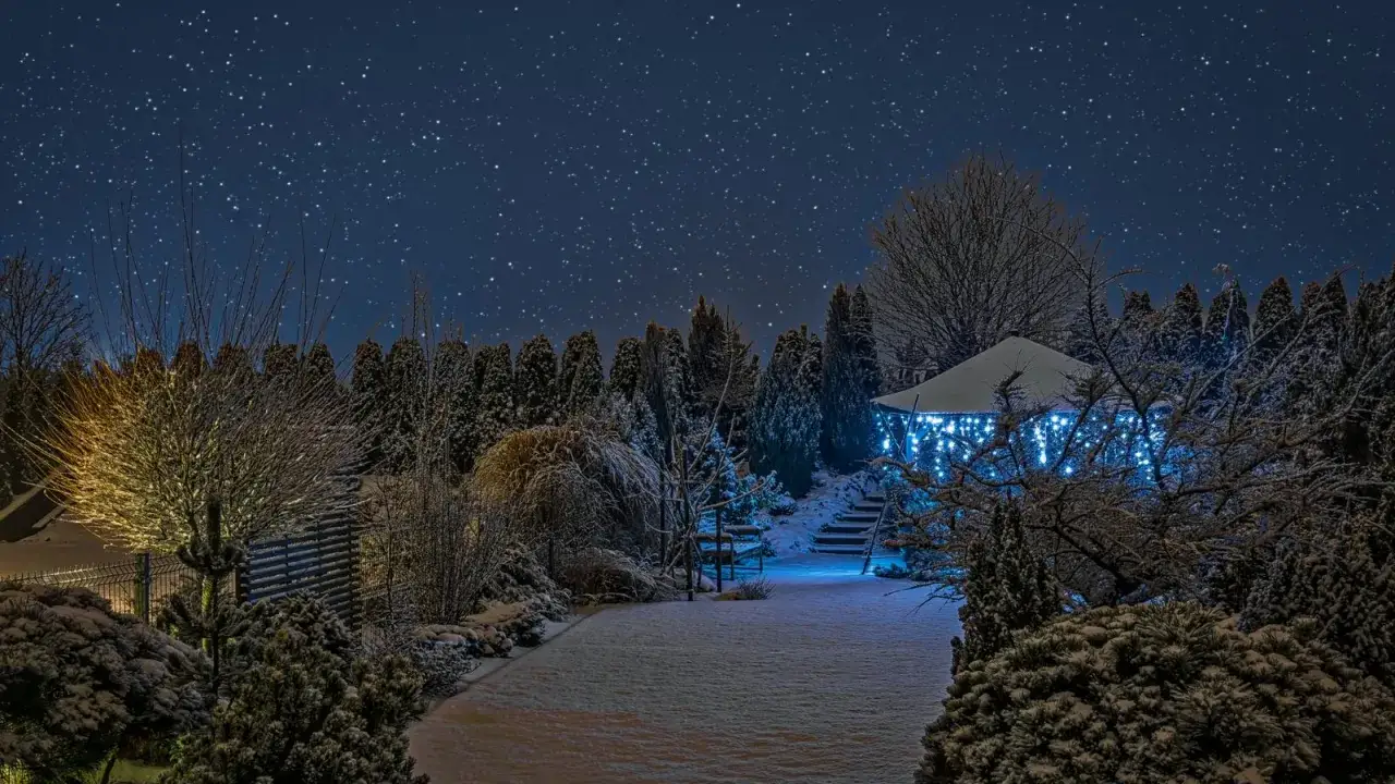 Ośnieżony ogród nocą, z altanką ozdobioną niebieskimi lampkami. Widać, że lampy solarne świecą zimą, oświetlając ścieżkę i drzewa.