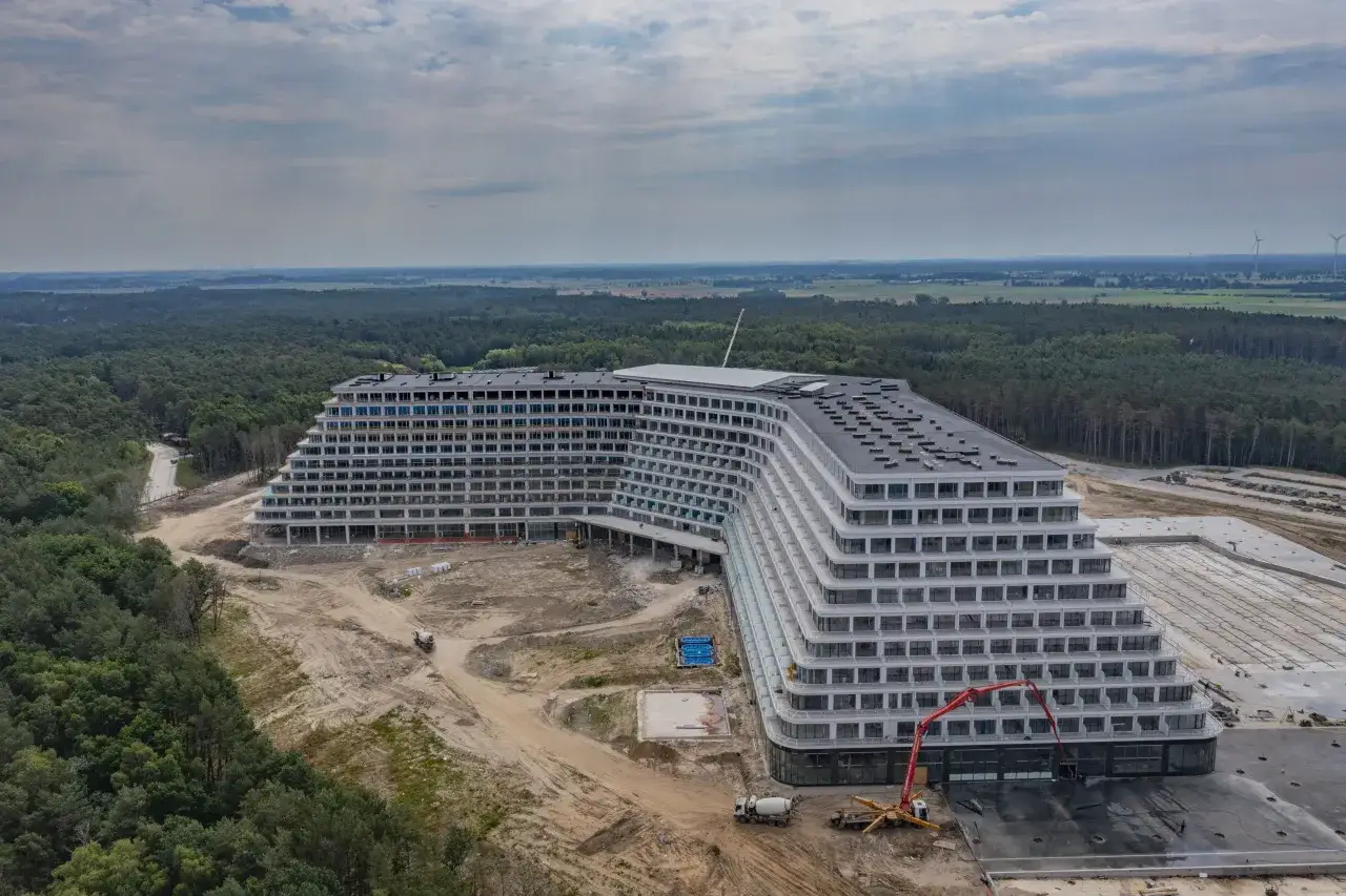 Ogromny, tarasowy hotel w budowie, który ma być największym hotelem nad morzem. Otoczony lasem, z widocznymi placem budowy i maszynami.