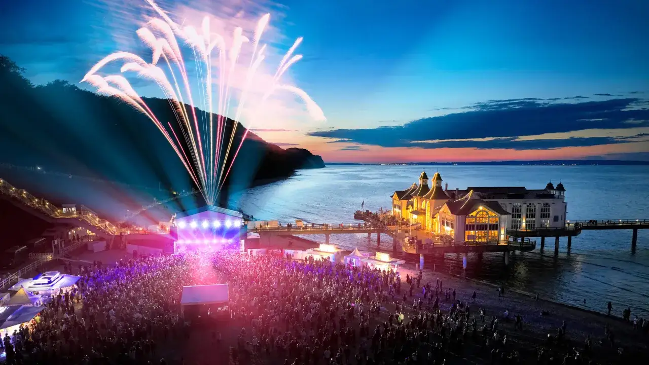 Feuerwerk über einem Strandkonzert an der Ostsee. Tausende feiern am Abend zu Live-Musik, während bunte Raketen den Himmel erhellen.