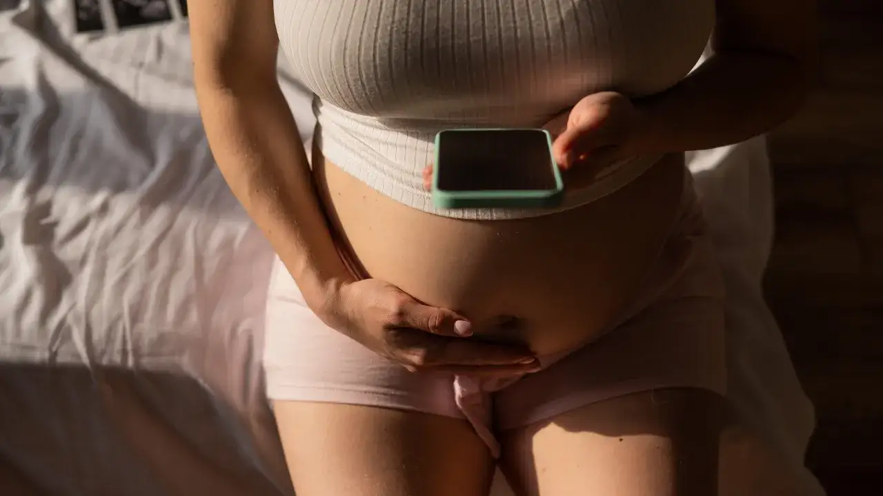 Kobieta w zaawansowanej ciąży, z dłońmi na twardym brzuchu przed porodem, trzyma telefon.