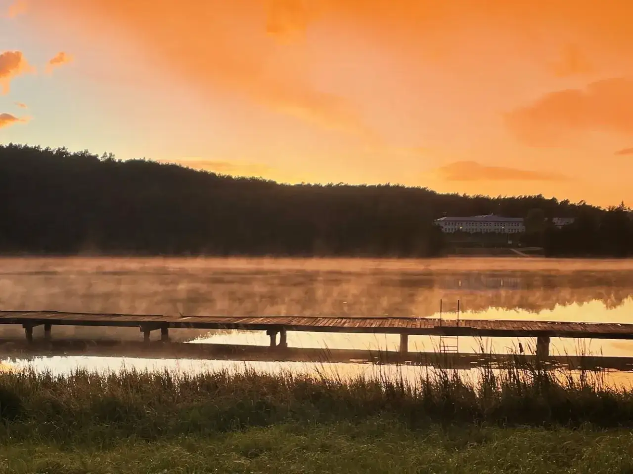 Mglisty poranek nad jeziorem w Torzymiu. Drewniane molo zanurza się w parującej wodzie, a na horyzoncie majaczy budynek.