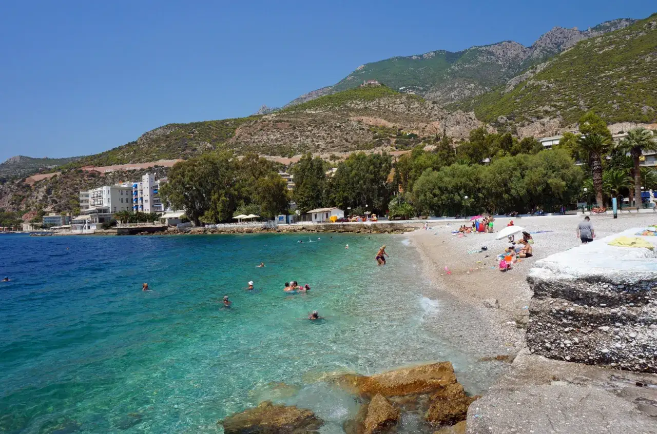 Słoneczny dzień na plaży w Loutraki, Grecja. Ludzie kąpią się w krystalicznie czystej wodzie, podczas gdy g&oacute;ry g&oacute;rują nad wybrzeżem.