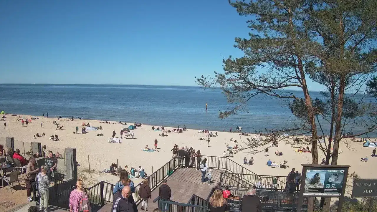 Słoneczna stegna plaża tętni życiem. Ludzie odpoczywają na piasku, kąpią się w morzu i spacerują po drewnianych schodach.