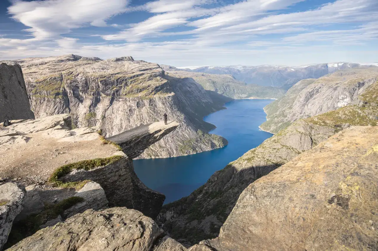 Widok na spektakularną trasę Trolltunga. Człowiek stoi na skalnej półce nad błękitnym jeziorem, otoczony surowymi, norweskimi krajobrazami.
