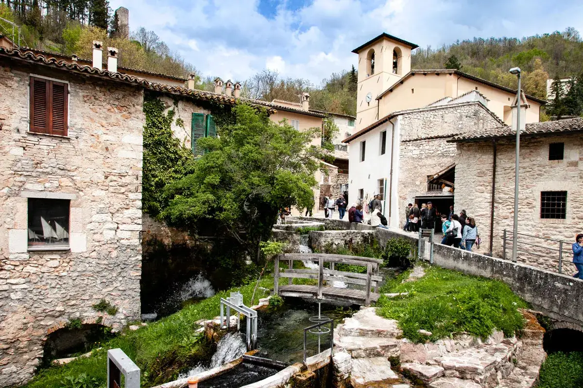 Un borgo pittoresco con piccole cascate e un ponte di legno. Le cascate del Menotre scorrono tra case in pietra, con persone che passeggiano.