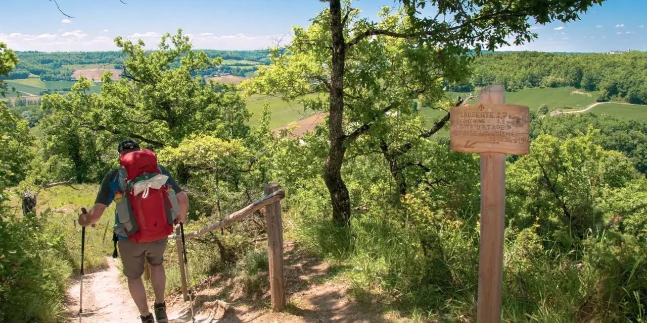Un randonneur avec un sac &agrave; dos rouge monte un sentier bois&eacute;, profitant des paysages verdoyants du Tarn-et-Garonne. Un panneau indique 