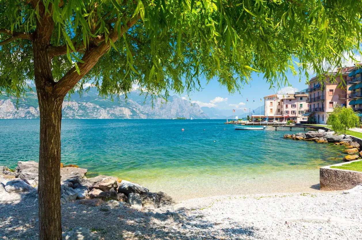 Un albero ombroso si affaccia su una spiaggia di ciottoli e acque cristalline, perfette per i **laghi balneabili**. In lontananza, montagne maestose e un villaggio colorato.
