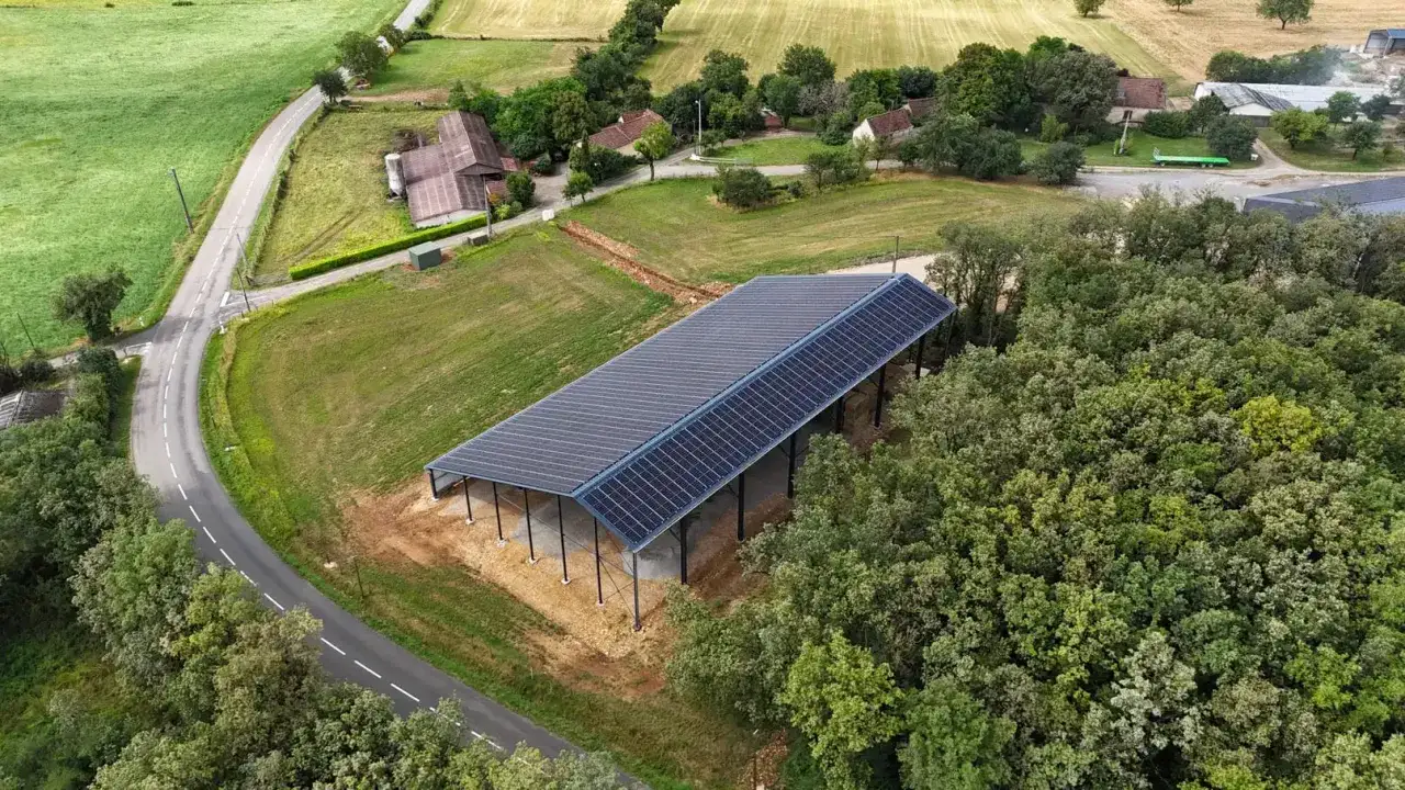 Vue aérienne d'un hangar agricole avec panneaux solaires sur le toit, entouré de verdure et de quelques habitations.
