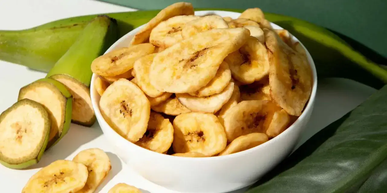 Schmackhafte, selbstgemachte Bananenchips in einer Schüssel, umgeben von frischen grünen Bananen. Ein gesunder Snack zum Selbermachen.
